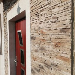 KAMIREM - Elewacja budynku wykończona kamieniem dekoracyjnym w odcieniach beżu i brązu, obok czerwone drzwi wejściowe z prostokątnymi przeszkleniami, całość otoczona jasną ramą.