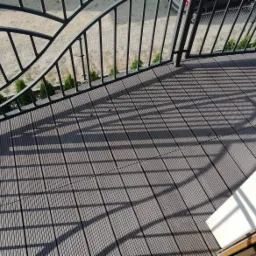 Szare deski kompozytowe na balkonie, cień barierki, widok z góry, rośliny w tle.