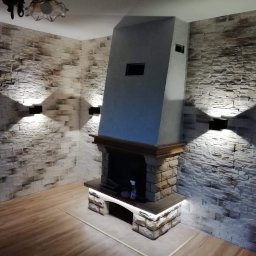 KAMIREM - Salon z kominkiem obłożonym kamieniem dekoracyjnym, oświetlonymi ścianami z imitacji kamienia i drewnianą podłogą.