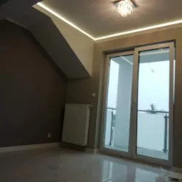 Jasne, błyszczące płytki podłogowe w pokoju na poddaszu z oknem balkonowym, grzejnikiem i nowoczesnym oświetleniem LED w suficie pod skosem.