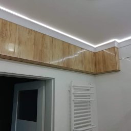 KAMIREM - Wykończenie sufitu w łazience z drewnopodobnymi panelami i oświetleniem LED, widoczny biały grzejnik drabinkowy i fragment kabiny prysznicowej.