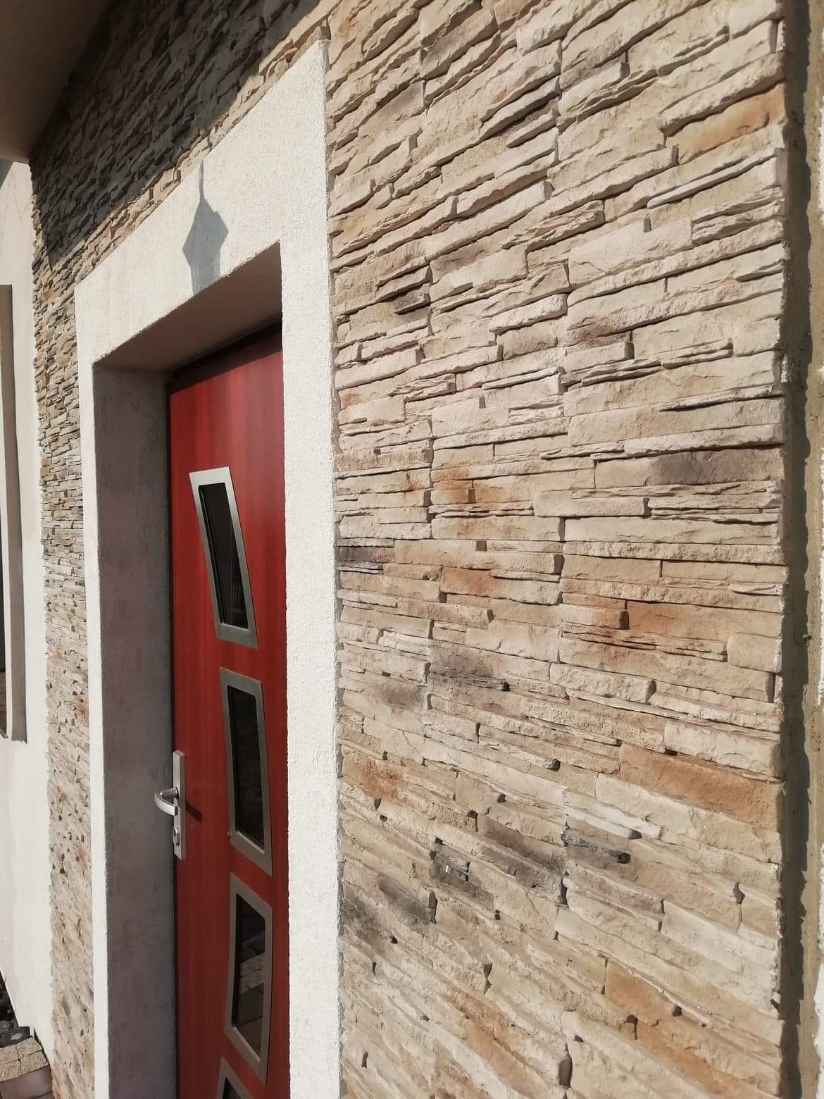 Elewacja budynku wykończona kamieniem dekoracyjnym w odcieniach beżu i brązu, obok czerwone drzwi wejściowe z prostokątnymi przeszkleniami, całość otoczona jasną ramą.
