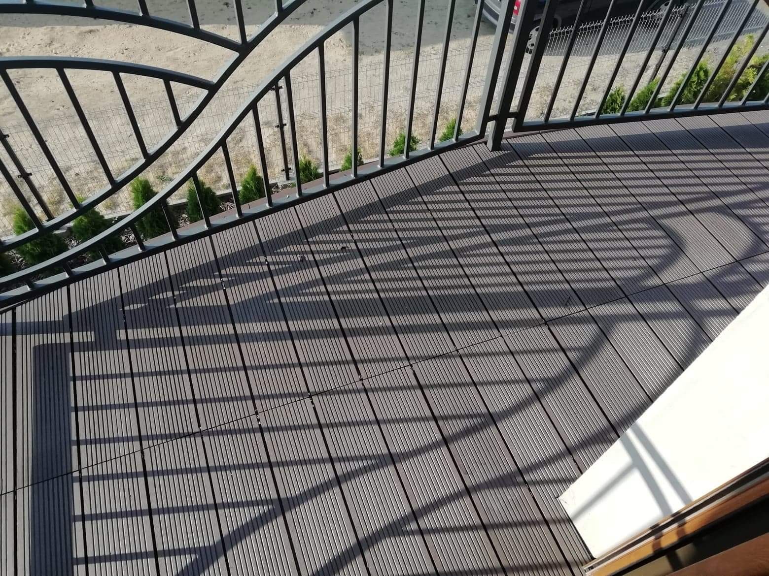 Szare deski kompozytowe na balkonie, cień barierki, widok z góry, rośliny w tle.