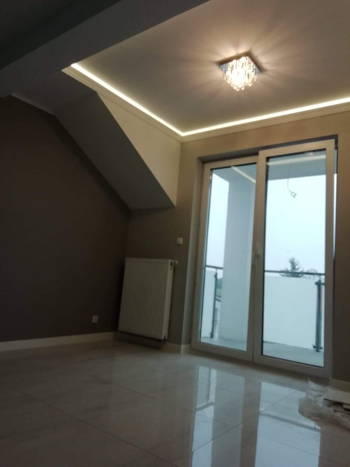 Jasne, błyszczące płytki podłogowe w pokoju na poddaszu z oknem balkonowym, grzejnikiem i nowoczesnym oświetleniem LED w suficie pod skosem.