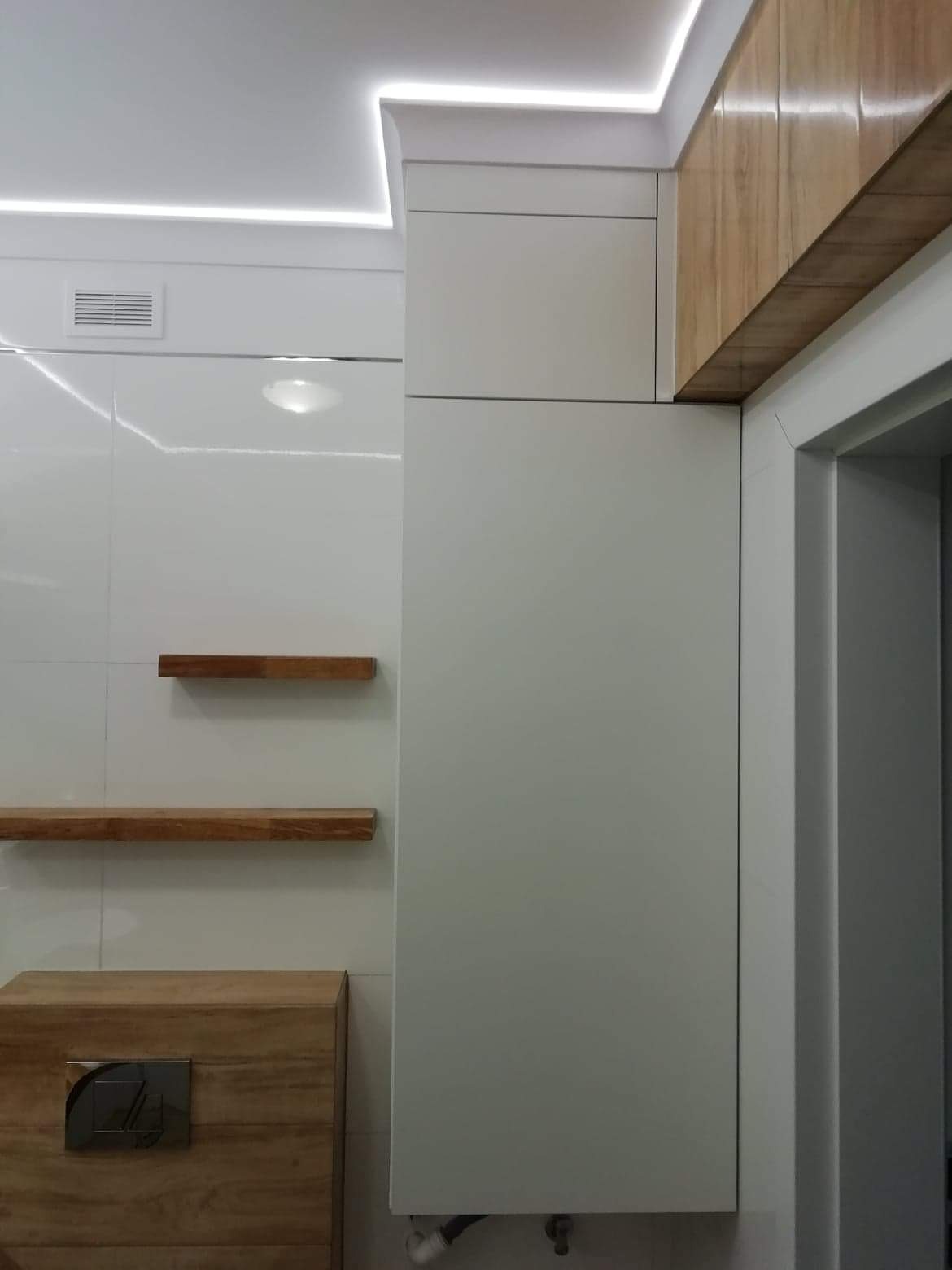 Nowoczesna łazienka z białymi, błyszczącymi płytkami, drewnianymi półkami i szafką pod umywalką z metalowym panelem sterującym spłuczką, oświetlenie LED podsufitowe.