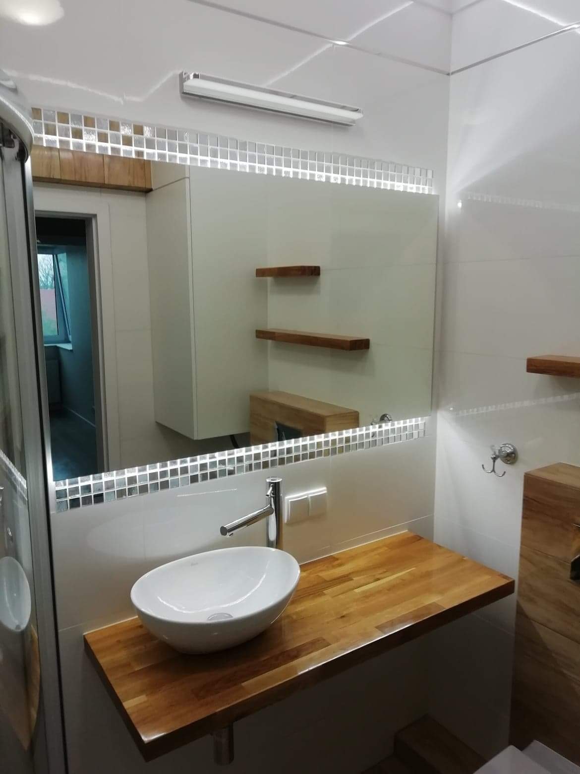 Biała umywalka nablatowa na drewnianym blacie w nowoczesnej łazience z podświetlanym lustrem obramowanym mozaiką i półkami z drewna.