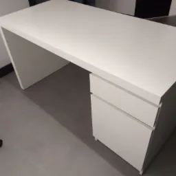 Nowoczesne, białe biurko z szufladami, ustawione na szarym tle, widoczne z perspektywy lekko z góry, z fragmentem krzesła biurowego w lewym dolnym rogu.