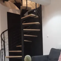 Nowoczesne schody spiralne z drewnianymi stopniami i czarną metalową konstrukcją, widoczne z perspektywy salonu z sofą i stolikiem kawowym z kaktusem.