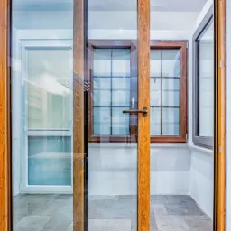 Przesuwne drzwi balkonowe z drewnianą ramą, widok na balkon z oknami szprosowanymi i roletą zwijaną.