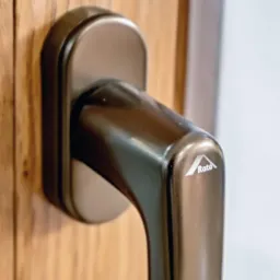 Brązowa klamka okienna z logo Roto zamontowana na drewnianej ramie okiennej, widoczna faktura drewna.