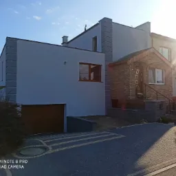 Dwukondygnacyjny dom z ocieploną elewacją w kolorze szarym, z widocznym garażem i wejściem wykończonym kamieniem elewacyjnym, oświetlony popołudniowym słońcem.