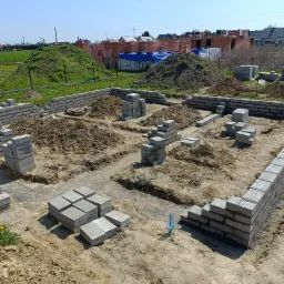 Wykopane fundamenty i częściowo wzniesione ściany fundamentowe z szarych bloczków betonowych na placu budowy w słoneczny dzień. W tle widoczne pryzmy cegieł i powstające budynki.