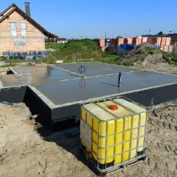 Wylany fundament pod budynek jednorodzinny, widoczny żółty zbiornik IBC na wodę, ściany fundamentowe pomalowane na czarno, w tle cegły i częściowo wzniesiony budynek.