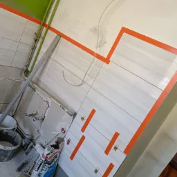 Układanie białych, poziomych płytek z pomarańczową taśmą wyznaczającą linie, widoczne narzędzia, poziomica, wiadro z zaprawą i worek kleju Ceresit.