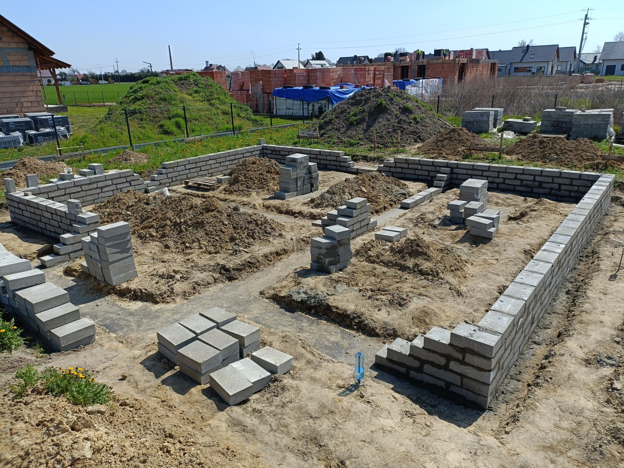 Wykopane fundamenty i częściowo wzniesione ściany fundamentowe z szarych bloczków betonowych na placu budowy w słoneczny dzień. W tle widoczne pryzmy cegieł i powstające budynki.