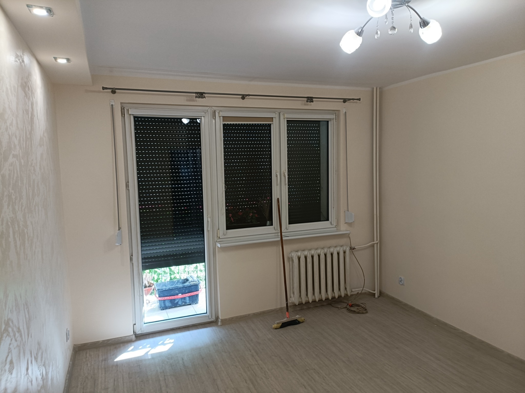 Pusta, jasna przestrzeń pokoju z oknem z roletami, grzejnikiem i śladami po remoncie: zmiotka oparta o ścianę, kabel na podłodze, widok na balkon z pojemnikiem.