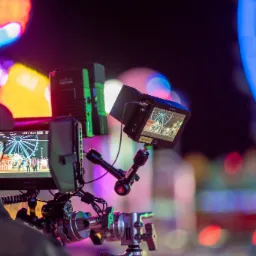 Kamera filmowa z podglądem na ekranie, rejestrująca scenę z karuzelą i ludźmi, ustawiona na statywie w scenerii nocnego parku rozrywki.