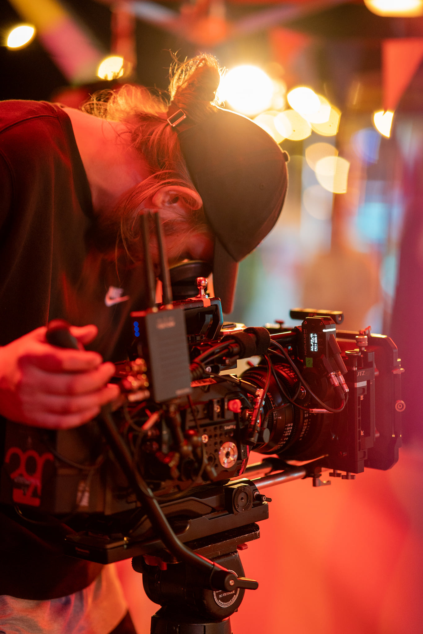 Operator z kucykiem w czapce, obsługujący profesjonalną kamerę filmową z monitorem i osprzętem, oświetloną czerwonym światłem na planie filmowym.