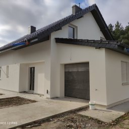 Aras bud - Nowoczesny, otynkowany na biało dom jednorodzinny w Bydgoszczy z ciemną dachówką, widoczny z poziomu gruntu, z wiadrem farby przy garażu.