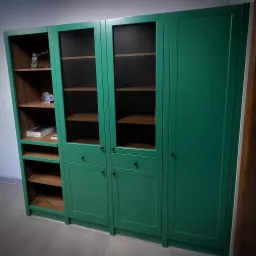 Zielona szafa na wymiar z drewnianymi półkami, przeszklonymi drzwiami i dolnymi szafkami. Widoczna zawartość półek i minimalistyczne uchwyty.