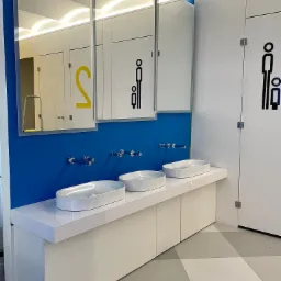 Nowoczesna łazienka publiczna z trzema umywalkami nablatowymi na białym blacie, błękitną ścianą i lustrami. Na drzwiach piktogramy.