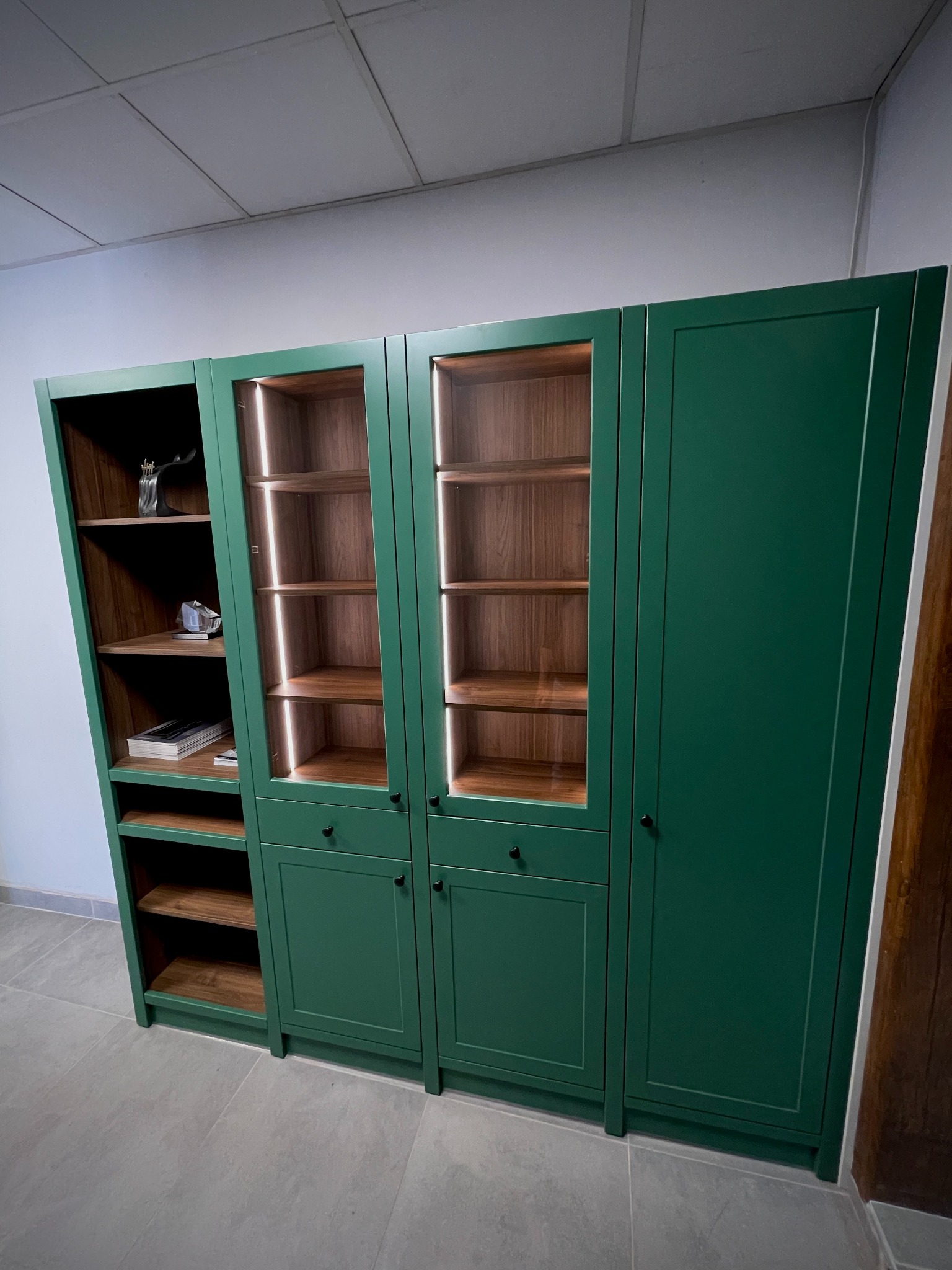 Regał z witrynami w odcieniach zieleni i drewna, oświetlony LED, wykonany na wymiar. Widoczne szklane fronty i drewniane półki. Aranżacja salonu.