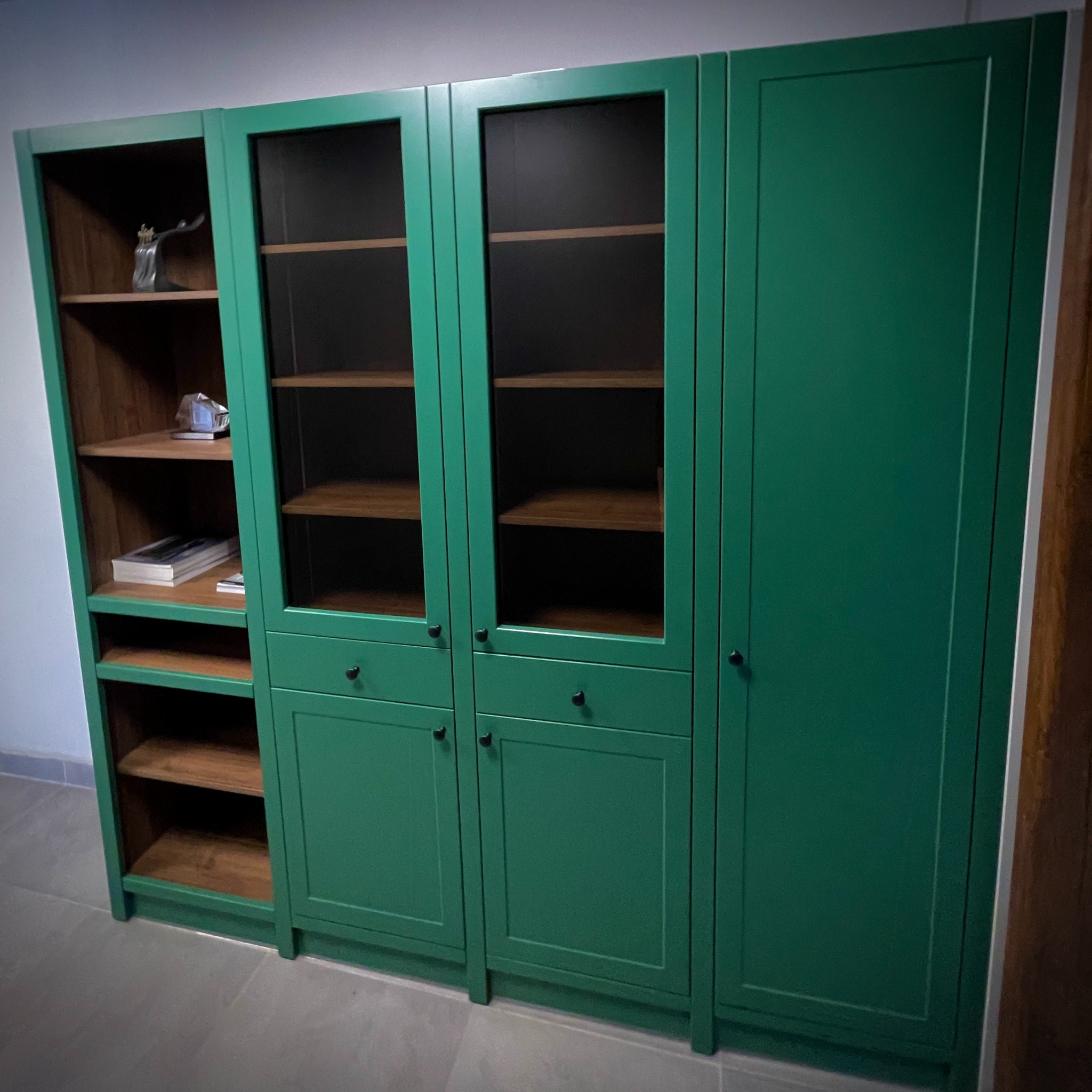 Zielona szafa na wymiar z drewnianymi półkami, przeszklonymi drzwiami i dolnymi szafkami. Widoczna zawartość półek i minimalistyczne uchwyty.