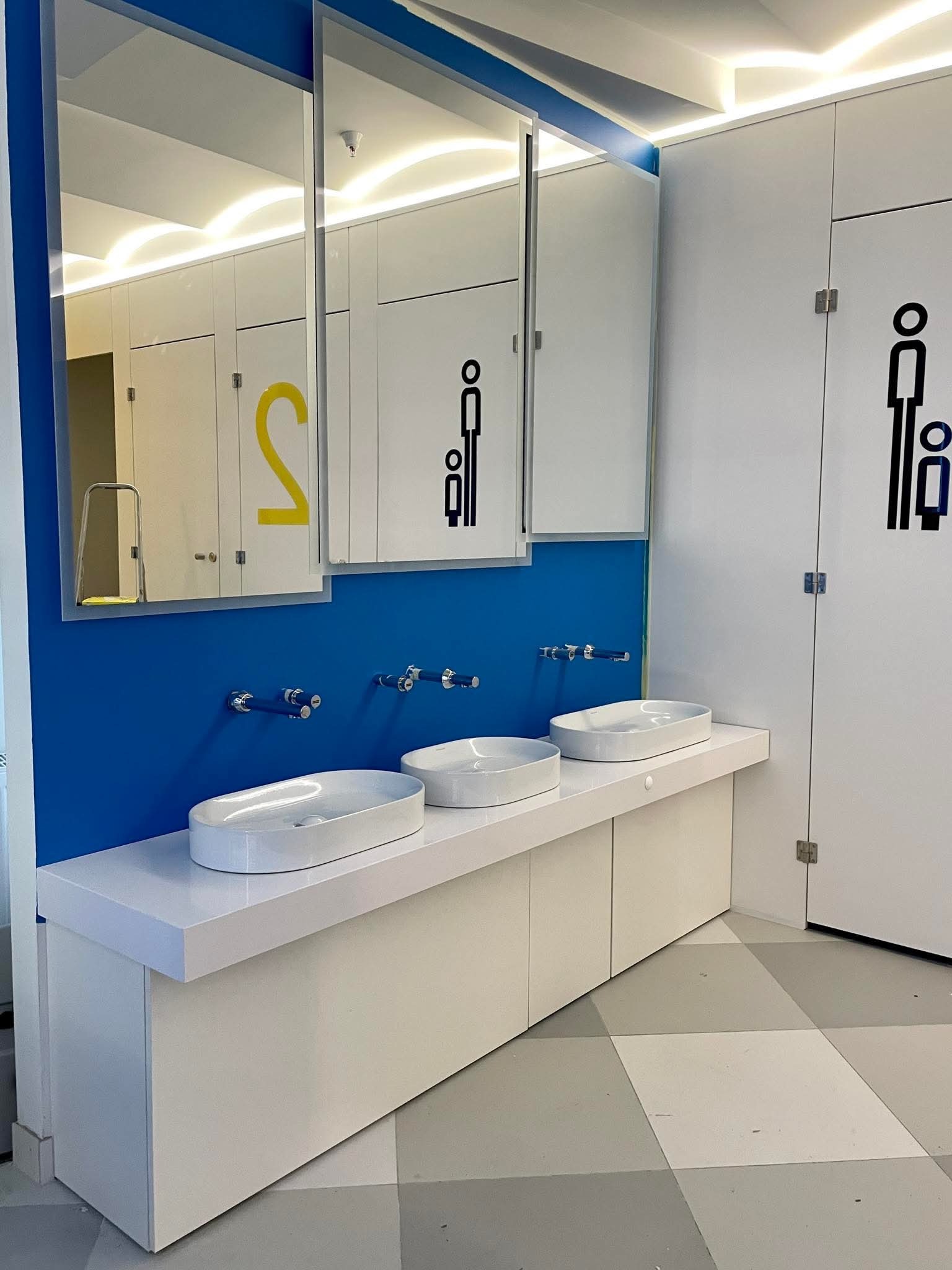 Nowoczesna łazienka publiczna z trzema umywalkami nablatowymi na białym blacie, błękitną ścianą i lustrami. Na drzwiach piktogramy.