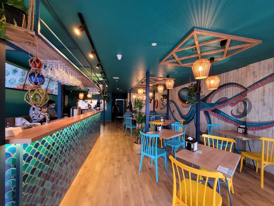 Wnętrze restauracji z motywem morskim: turkusowy sufit, bar z płytkami w kształcie rybich łusek, mural z ośmiornicą na ścianie, drewniane stoły i kolorowe krzesła.