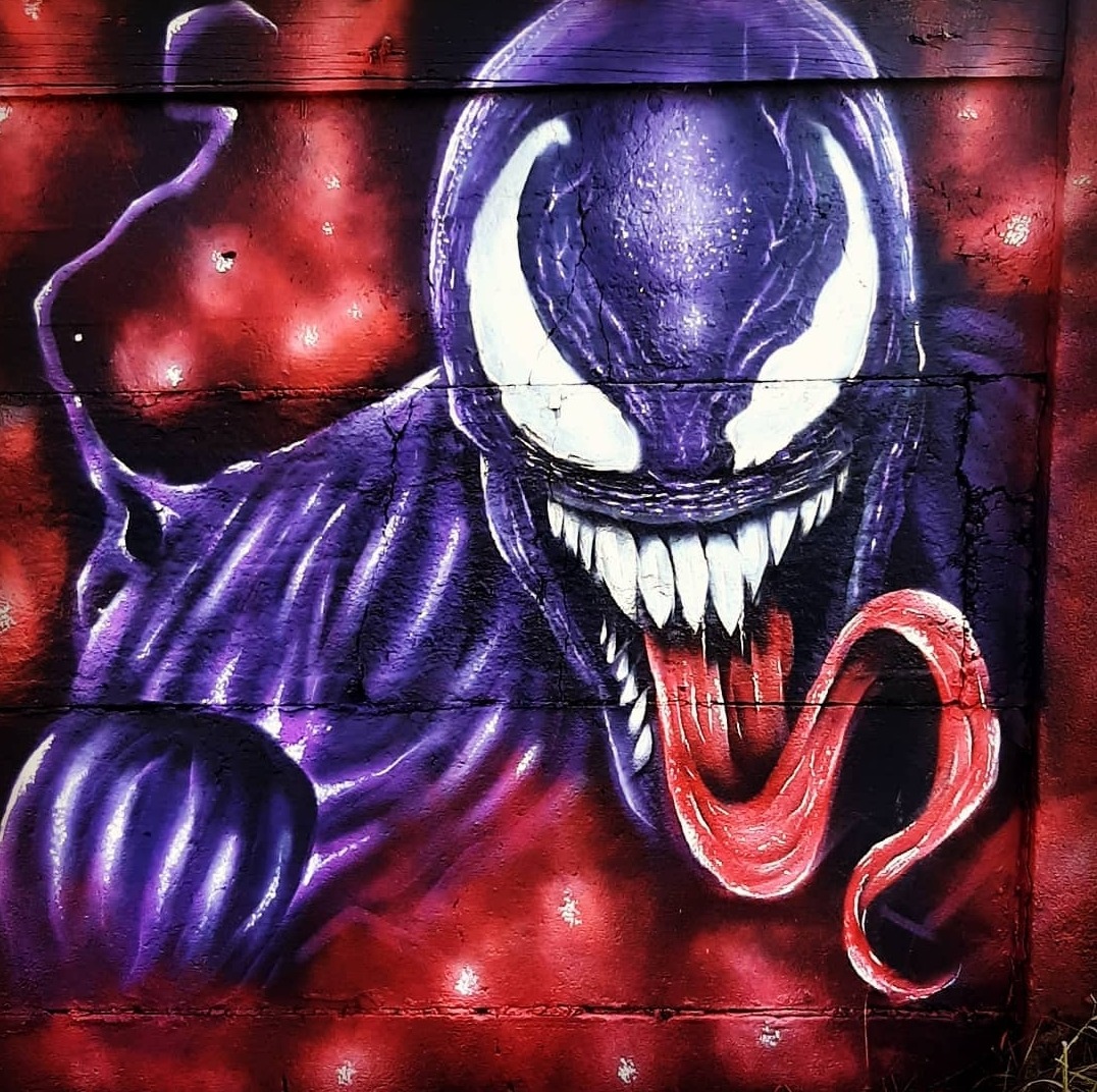 Fioletowo-czerwony mural na drewnianej ścianie przedstawiający postać Venom z komiksów Marvela, z detalami pęknięć w farbie i widoczną strukturą drewna.