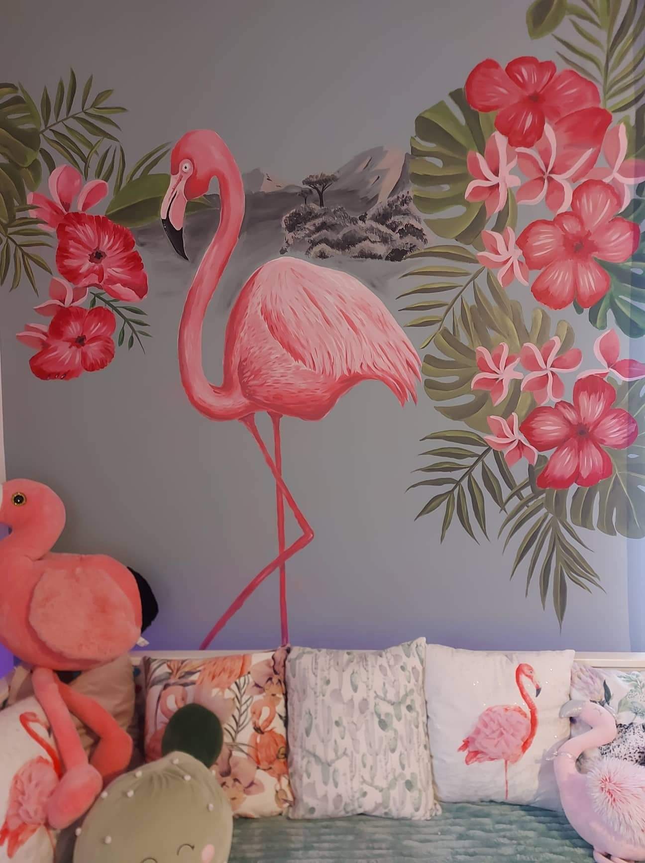Ręcznie malowany flaming na ścianie w pokoju dziecięcym, otoczony tropikalnymi kwiatami i liśćmi, z widocznymi poduszkami i pluszowym flamingiem na łóżku.