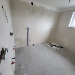 Surowe wnętrze pokoju w trakcie remontu: ściany w szpachli, wystające kable elektryczne, rury hydrauliczne, surowa wylewka na podłodze i fragment rozkopanej podłogi w rogu.