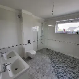 Jasna łazienka z podwójną umywalką, kabiną prysznicową typu walk-in i wiszącą toaletą. Na podłodze widoczna kombinacja szarych płytek z wzorem i bez. Z sufitu zwisa niepodłączona żarówka.