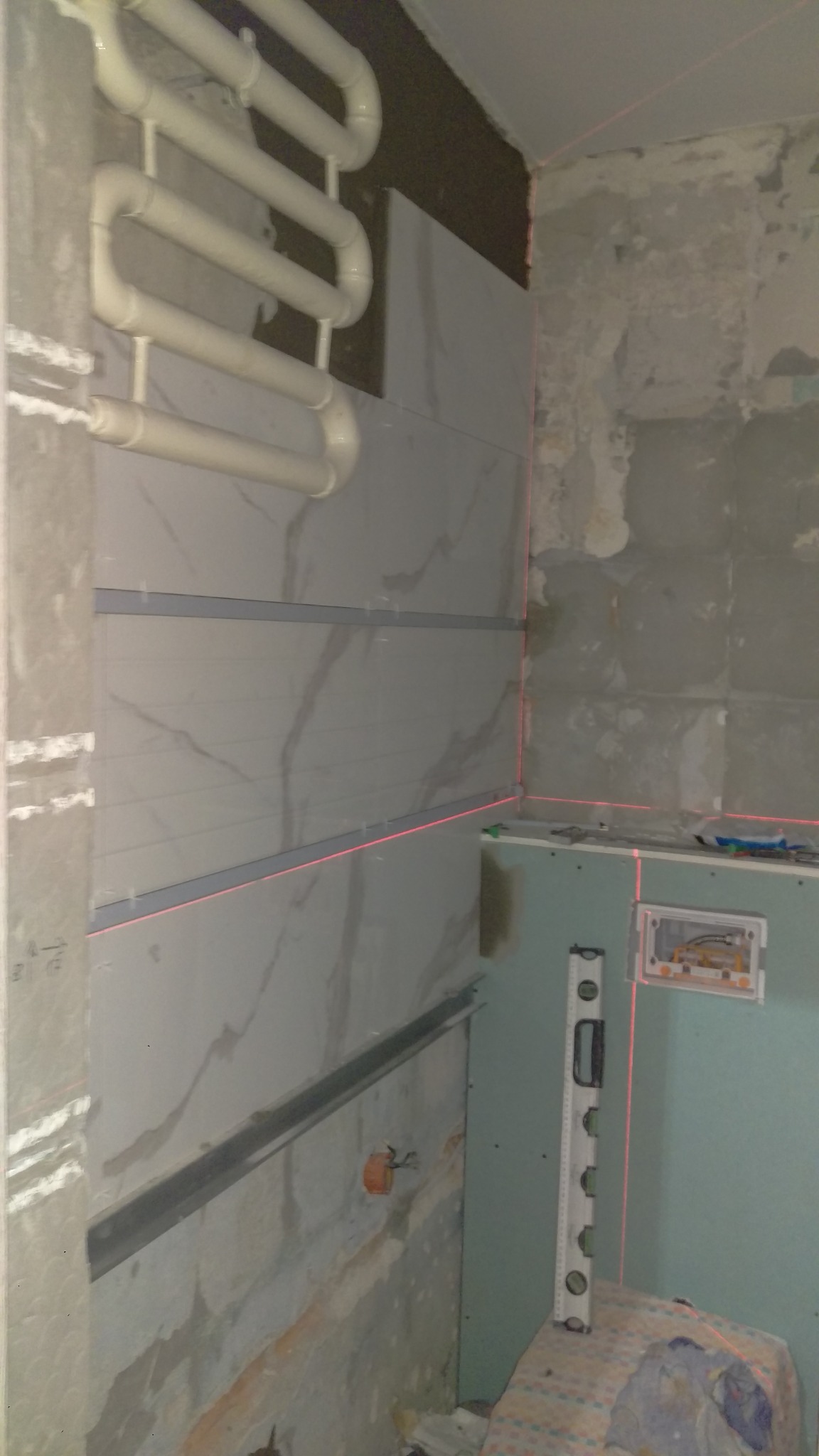 Montaż płytek imitujących marmur w łazience, widoczny grzejnik drabinkowy, profil montażowy i poziomica laserowa, ściany w stanie surowym.