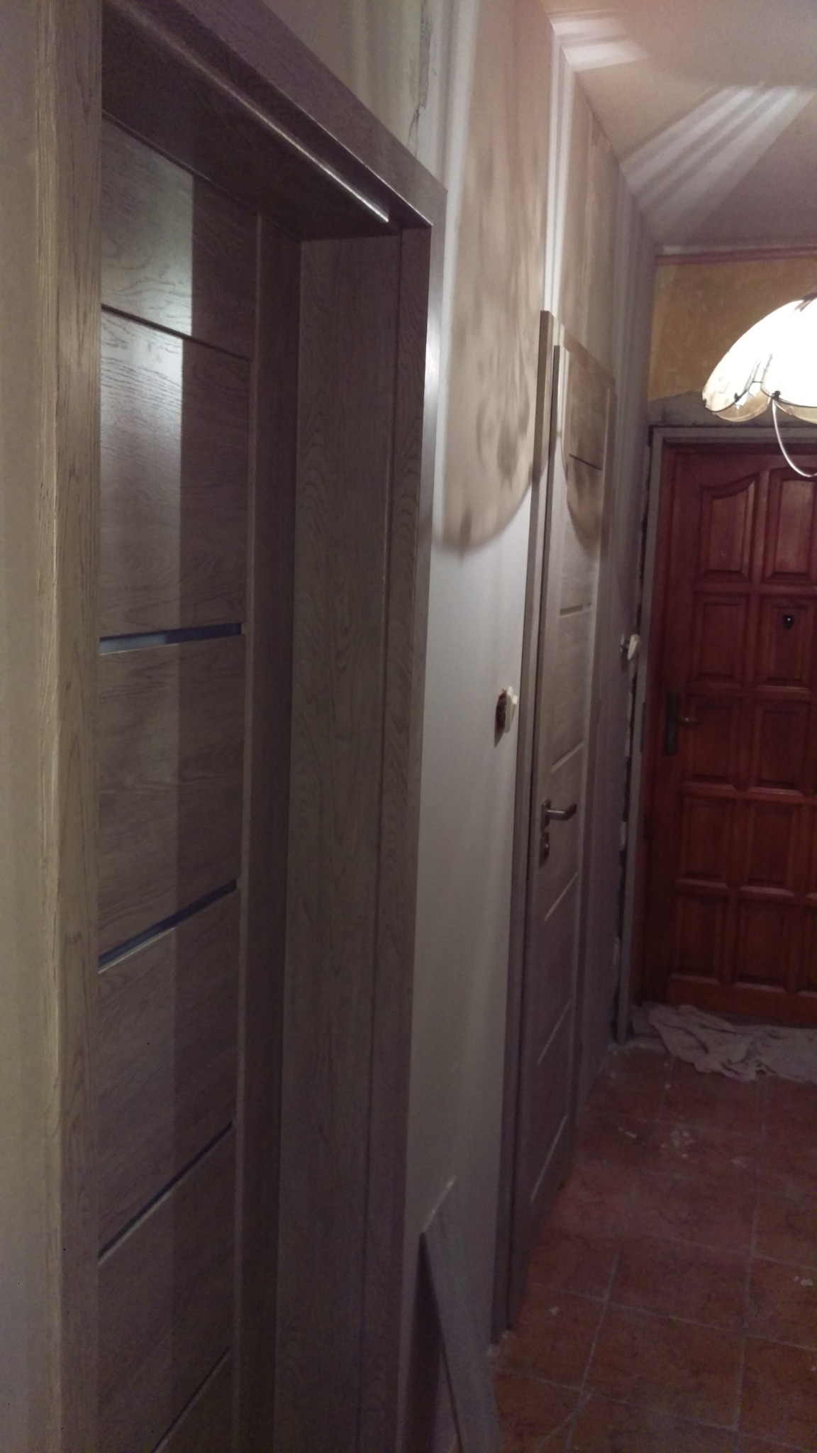 Montaż wewnętrznych drzwi z drewnianą okleiną i poziomymi wstawkami w trakcie remontu mieszkania, widoczne niedoskonałości na ścianie i podłodze.