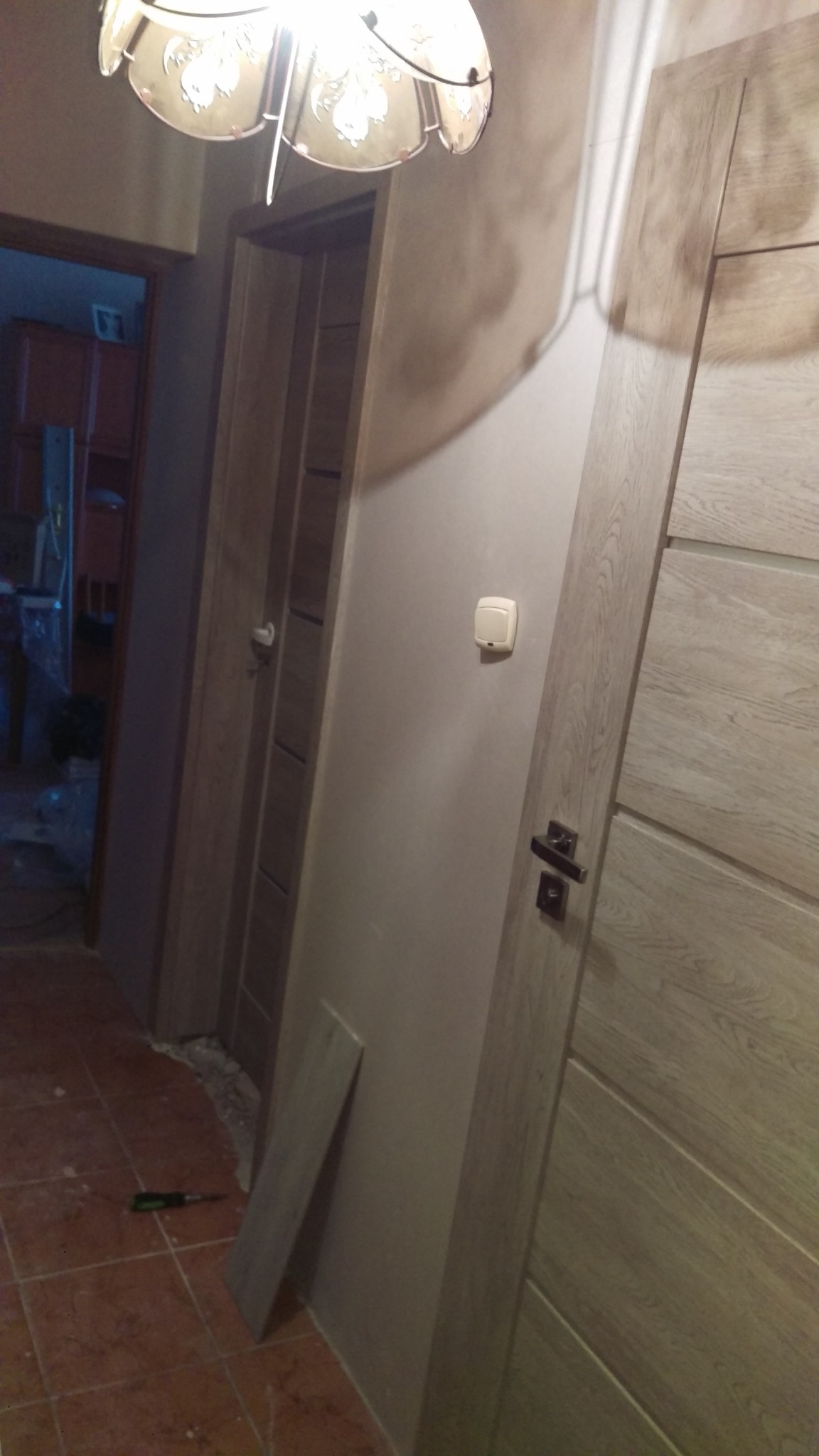 Wnętrze mieszkania w trakcie wykańczania: dwie drewniane, jasne drzwi, ściana z włącznikiem, fragment podłogi z terakoty, narzędzie i płyta przy ścianie.