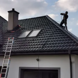 Pracownik w uprzęży, z myjką ciśnieniową na dachu z ciemnej dachówki, czyszczący dach z zanieczyszczeń. Widoczny komin, okna dachowe, drabina oparta o budynek i fragment elewacji.
