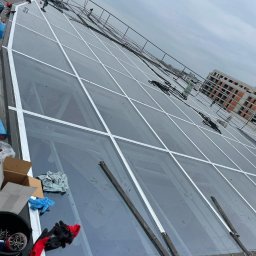 AlpinKris Krzysztof Wierzbowicz - Szklany dach z białymi ramami w trakcie budowy lub naprawy, widoczne narzędzia i rękawice robocze, budynek w tle.