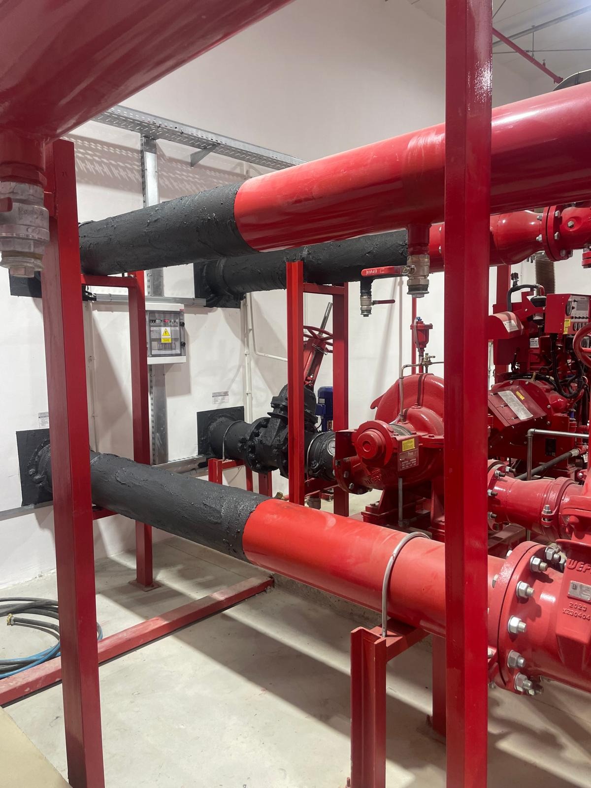 System rur ppoż. z czerwonymi rurami i czarną izolacją, widoczne zawory, pompy i okablowanie w jasnym pomieszczeniu technicznym z białą ścianą i szarą podłogą.