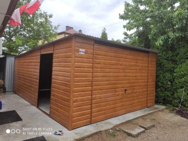 Brązowy garaż z blachy trapezowej, z częściowo otwartymi drzwiami, na betonowej wylewce, widok z boku, flaga Polski w tle.