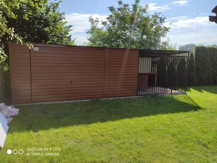 Brązowy garaż blaszany z dobudowaną klatką dla psa, postawiony na trawniku w ogrodzie, widok z boku.