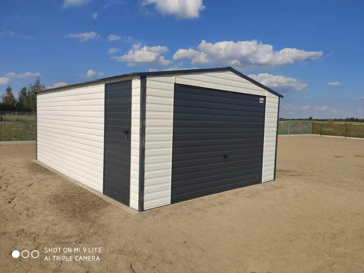 Gotowy garaż blaszany z bramą segmentową w kolorze grafitowym i białymi ścianami, ustawiony na wyrównanym terenie z widocznym ogrodzeniem w tle, sfotografowany w słoneczny dzień.