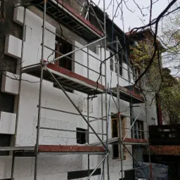 Budynek w trakcie ocieplania elewacji, widoczne rusztowania, płyty styropianowe częściowo zamontowane, resztki materiał&oacute;w budowlanych na ziemi.