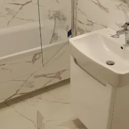 Nowoczesna łazienka z białą wanną za szklanym panelem i umywalką na szafce, wykończona płytkami imitującymi marmur.