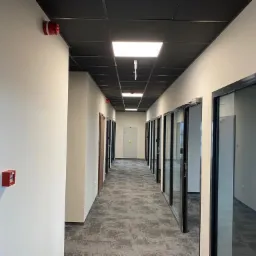 Długi korytarz biurowy z białymi ścianami, czarnym sufitem kasetonowym, szarą wykładziną dywanową i przeszklonymi drzwiami do biur po prawej stronie, widoczne czujniki dymu i przyciski alarmowe.