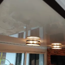 Błyszczący sufit z widocznymi odbiciami elementów wystroju wnętrza i dwóch nowoczesnych lamp wiszących z ażurowymi kloszami, na tle ceglanej ściany.