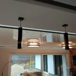 Nowoczesny sufit podwieszany z oświetleniem LED, lampy wiszące z abażurami, ściana z cegły dekoracyjnej w salonie z dużym oknem przesuwnym.