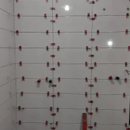 Świeżo ułożone, białe płytki ścienne w łazience, zabezpieczone czerwonymi klipsami poziomującymi i krzyżykami dystansowymi, widoczna poziomica przy ścianie i bałagan na podłodze.