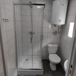 Wykończona łazienka z kabiną prysznicową ze szklanymi drzwiami, toaletą, bojlerem elektrycznym i szarymi płytkami podłogowymi oraz białymi płytkami ściennymi