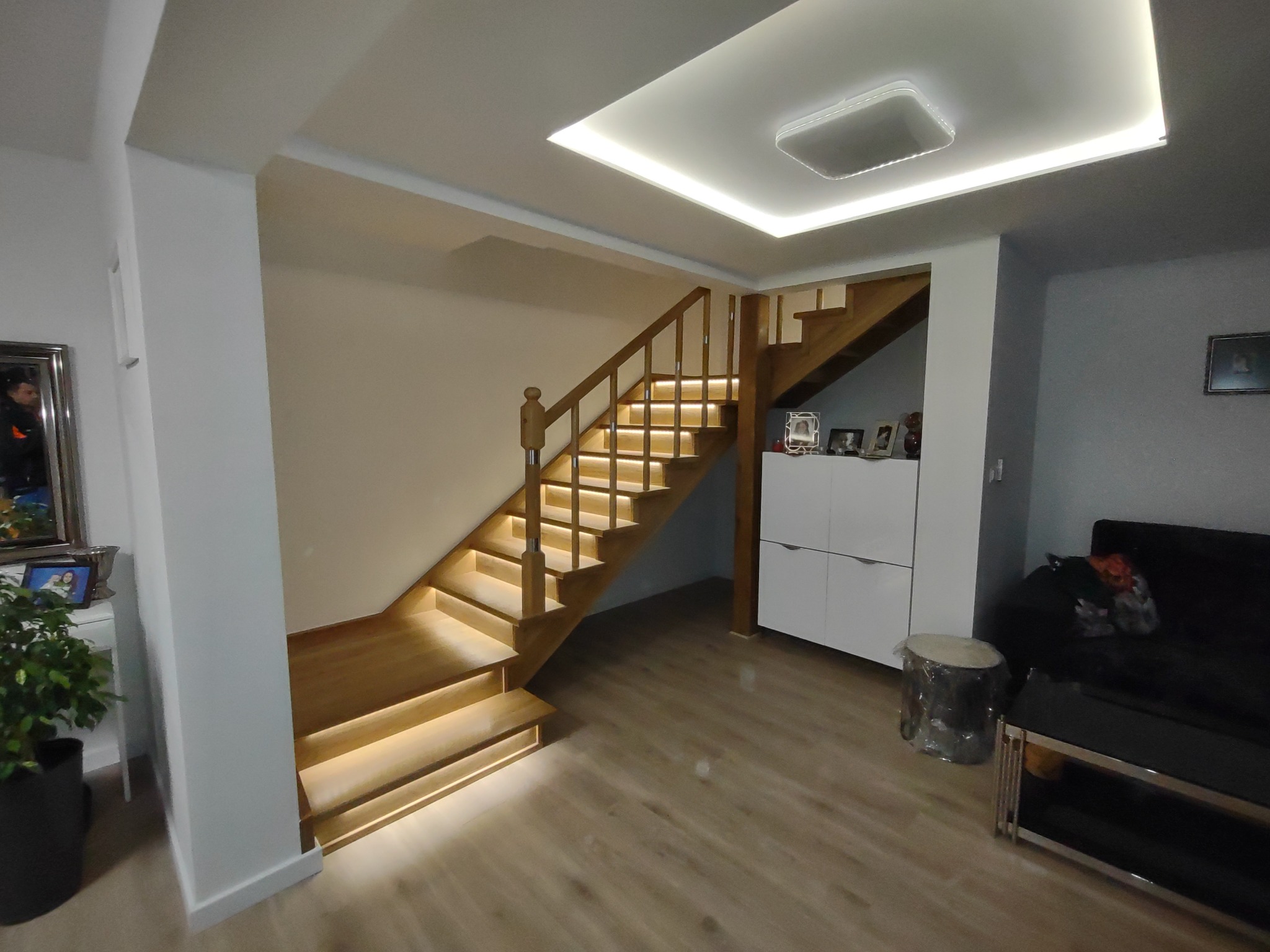 Drewniane schody z podświetleniem LED w nowoczesnym salonie z białymi ścianami i drewnianą podłogą, widoczne oświetlenie sufitowe i fragment czarnej sofy.